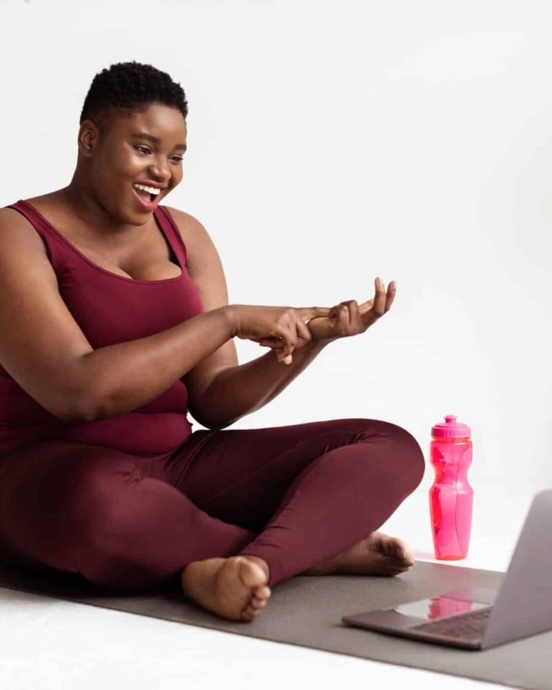 Cheerful plump millennial black woman with short hair yoga coach streaming from fitness studio, sitting on yoga mat with laptop and bottle of water, having video call with her students, copy space