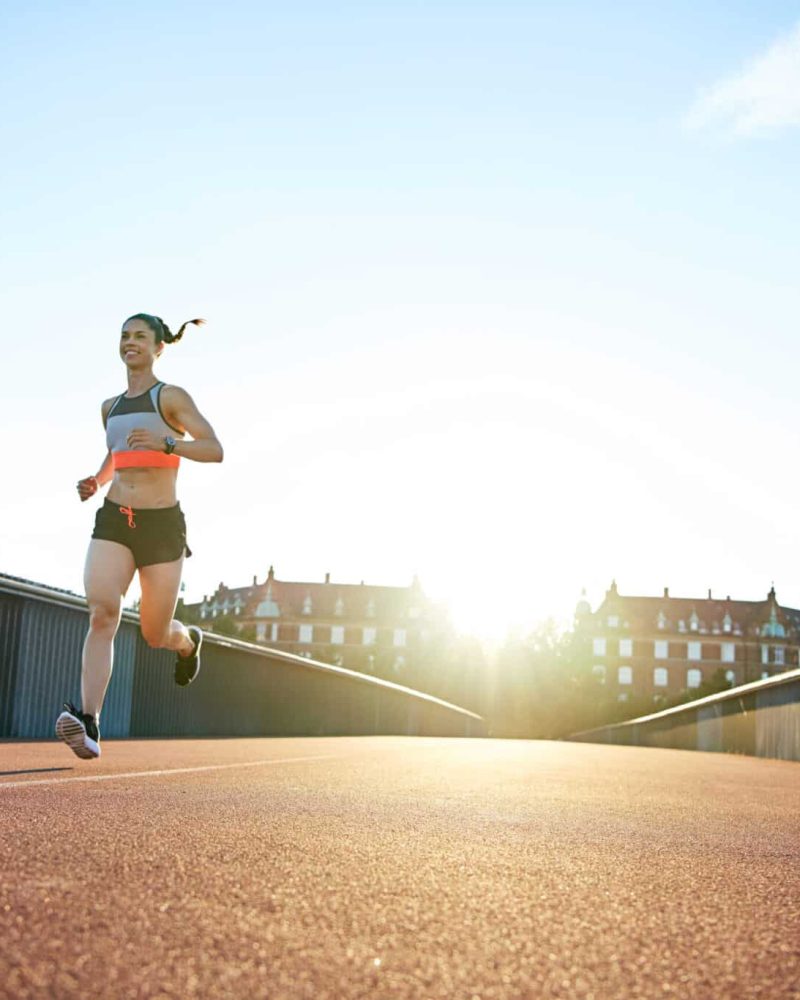 Low angle view of woman running down concrete and iron railing bridge with bright sunlight reaching over urban theme background