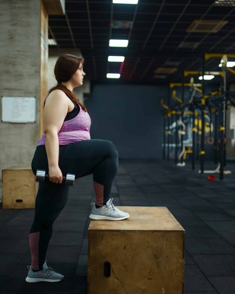 Overweight woman poses with dumbbells in gym, side view, active training. Obese female person struggles with excess weight, aerobic workout against obesity, sport club