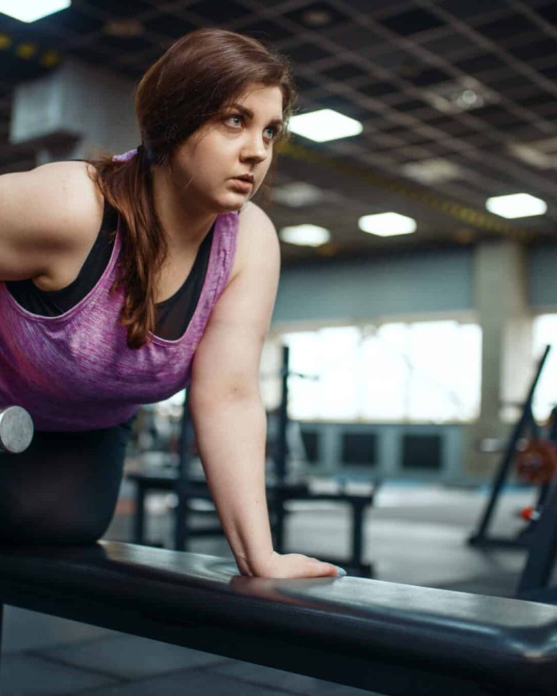 Overweight woman poses with dumbbells on brench in gym, active training. Obese female person struggles with excess weight, aerobic workout against obesity, sport club